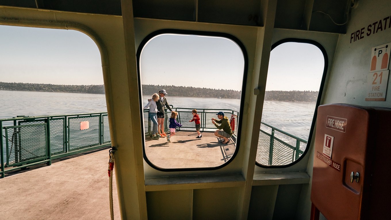A family stands and plays on the open deck of a ferry, viewed through rectangular windows from inside the vessel. Calm water and a distant treeline stretch across the background under a clear sky. Warm sunlight casts long shadows, giving the scene a relaxed, vacation-like atmosphere.